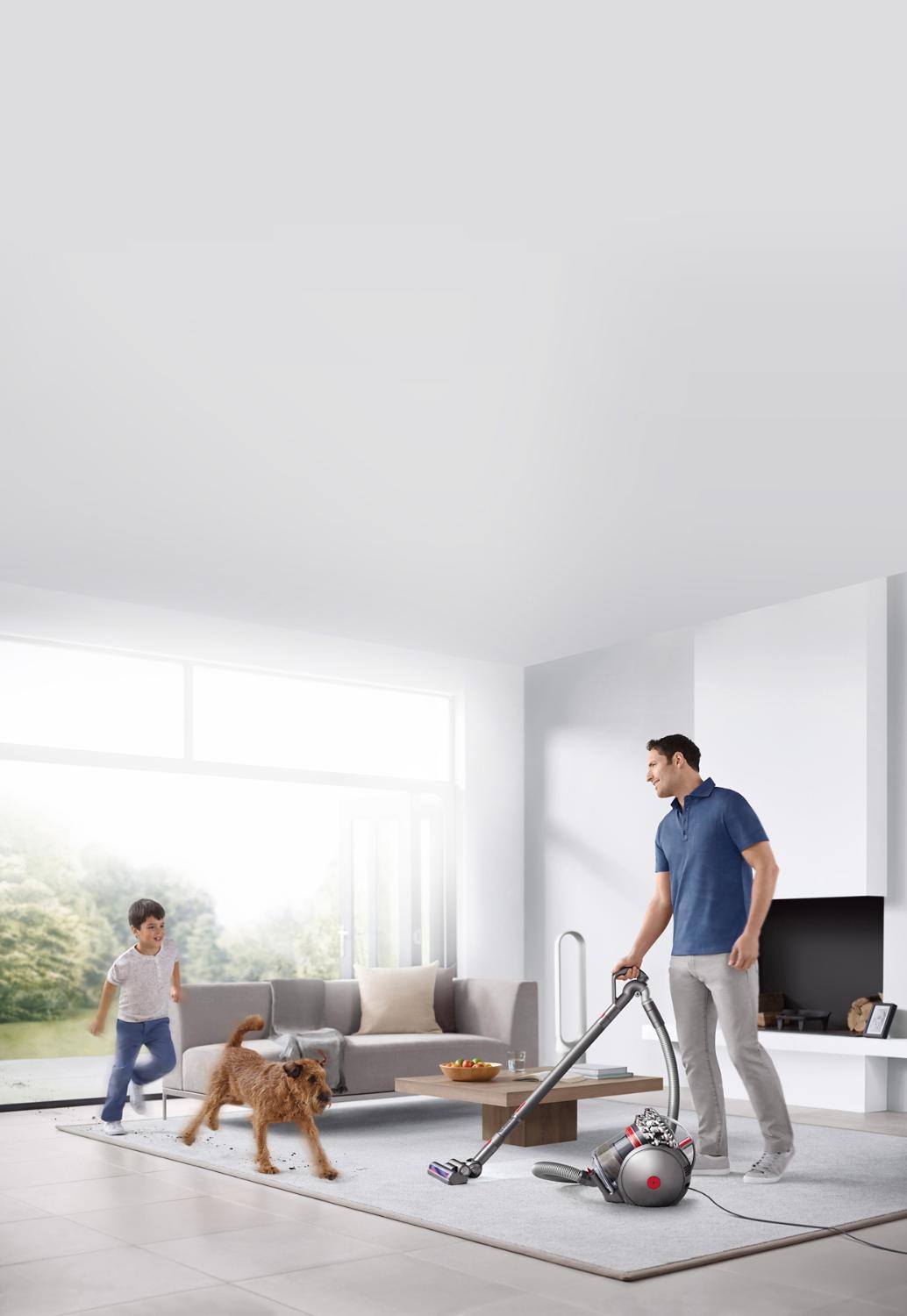 Un homme passant l’aspirateur dans la chambre en présence d’un chien et d’un enfant.