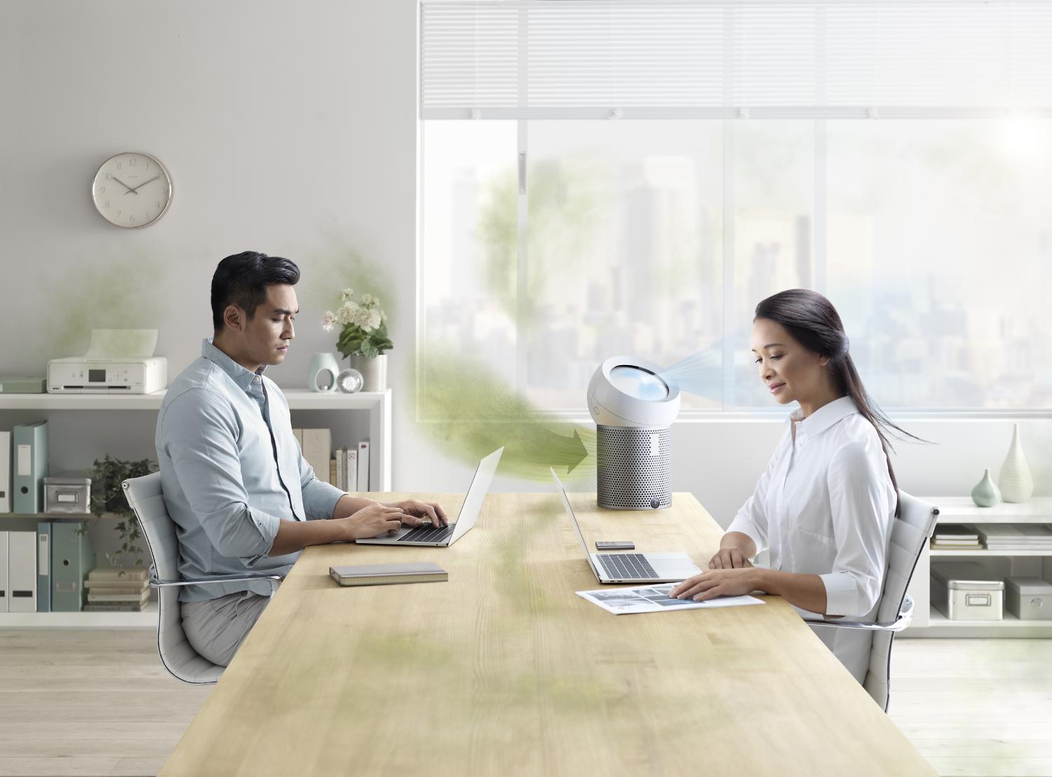 Two people working at a desk with the Dyson Pure Cool Me personal purifying fan injesting dirty air and projecting purfied air.