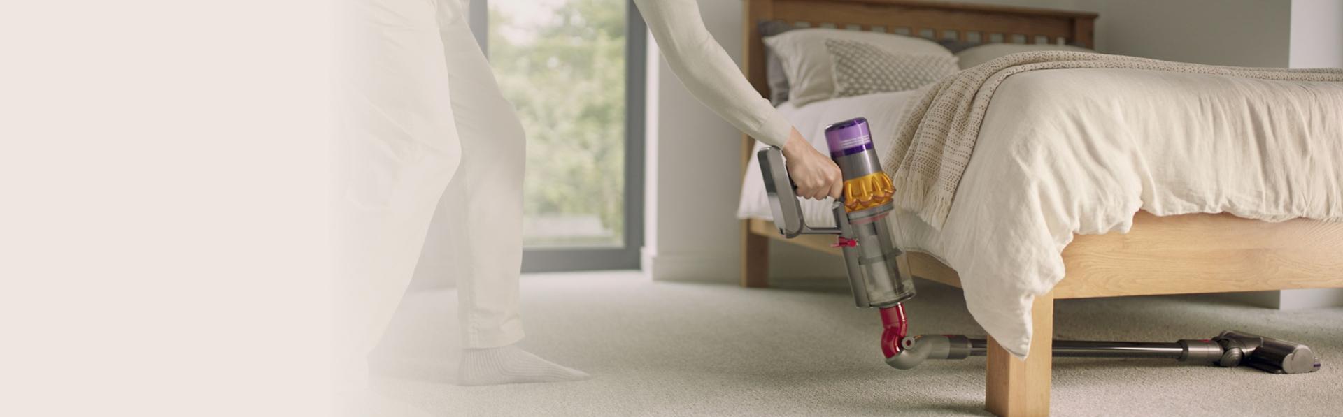 A person cleans deep underneath a bedframe with a Dyson vacuum and the Low-reach adaptor.