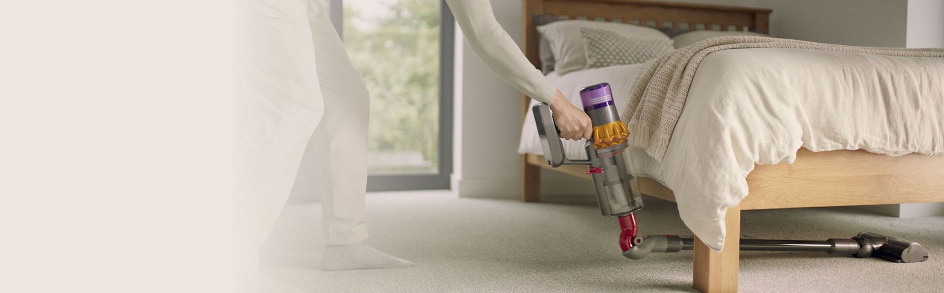  A person cleans deep underneath a bedframe with a Dyson vacuum and the Low-reach adaptor.