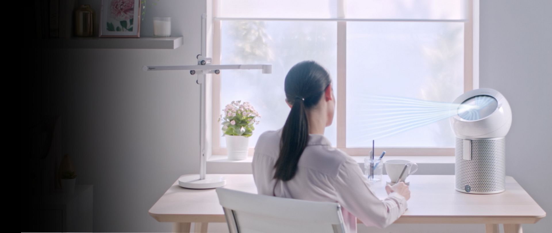 A woman working at a desk in her home while using a Dyson Pure Cool Me purifying fan