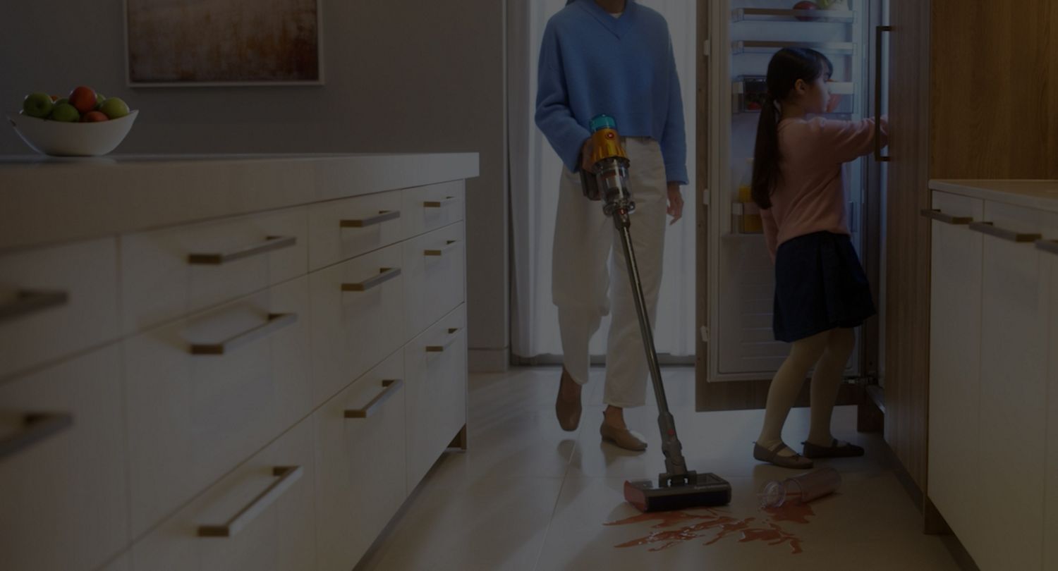 A child getting food from the fridge and spilling some. A parent cleans up the mess using a Dyson cordless vacuum