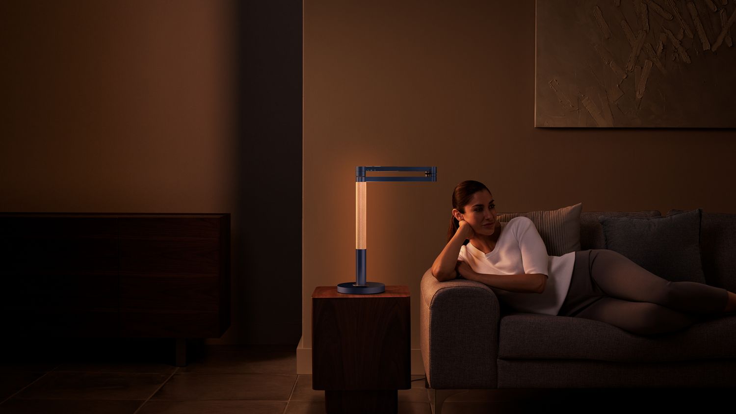 Une femme se détend sur le canapé avec la lampe Dyson Solarcycle Morph face à sa tige perforée, créant une lueur et une ambiance chaleureuses.
