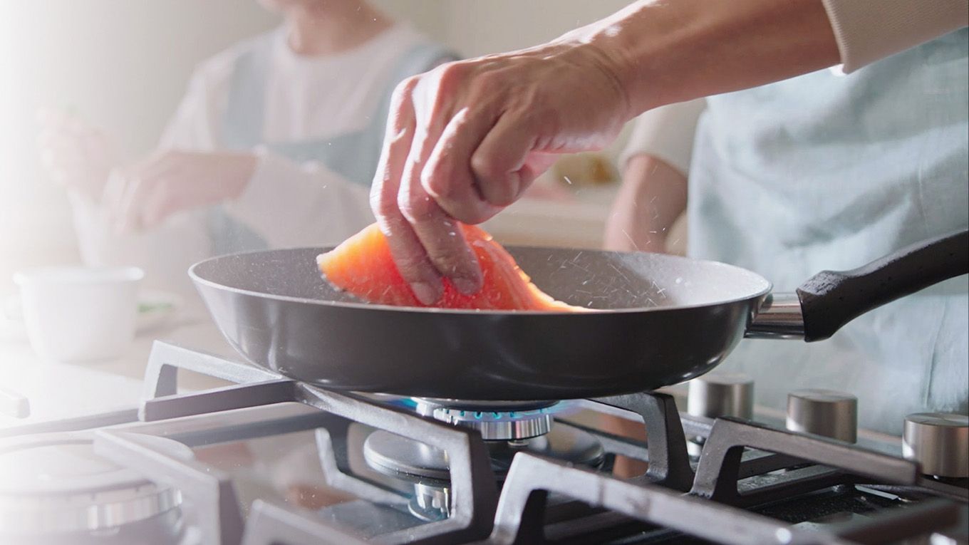 A chef lays a salmon fillet into a hot frying pan over a gas stove.