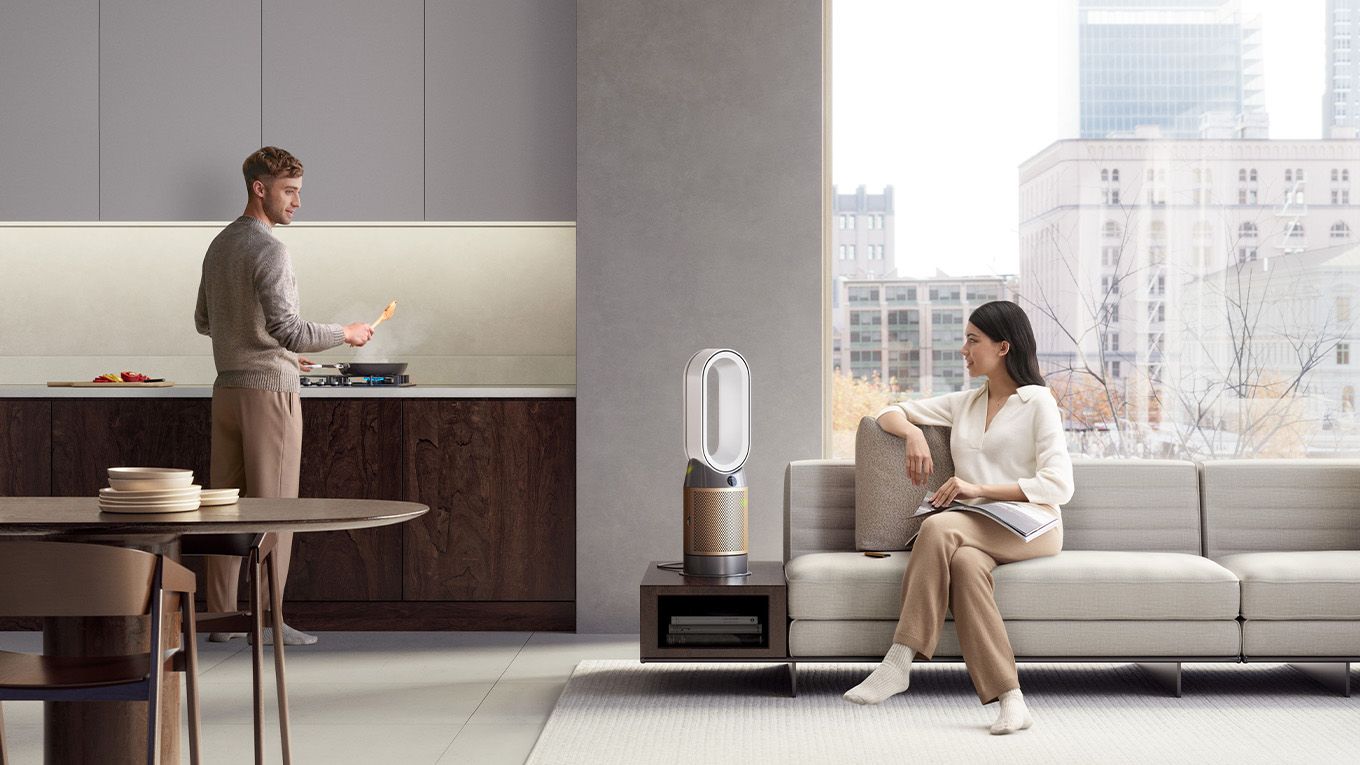 A man is cooking a meal on a stovetop in an apartment, a Dyson purifier cleans pollutants from the air and stops them reaching a woman who is sat on a sofa.