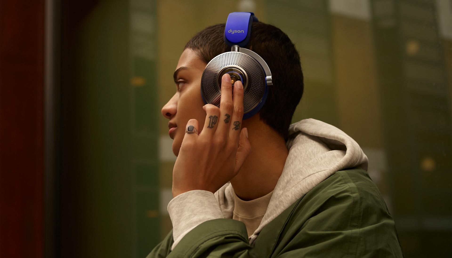 A model sitting against a window in the city while wearing her Dyson Zone noise cancelling headphones and tapping on the cup.