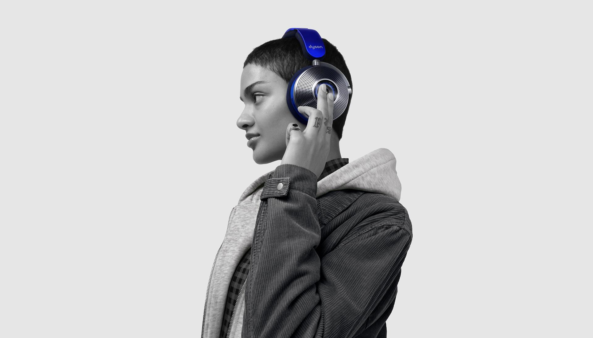A black and white picture of a model tapping the cup on their Dyson Zone noise cancelling headphones while against a white background.