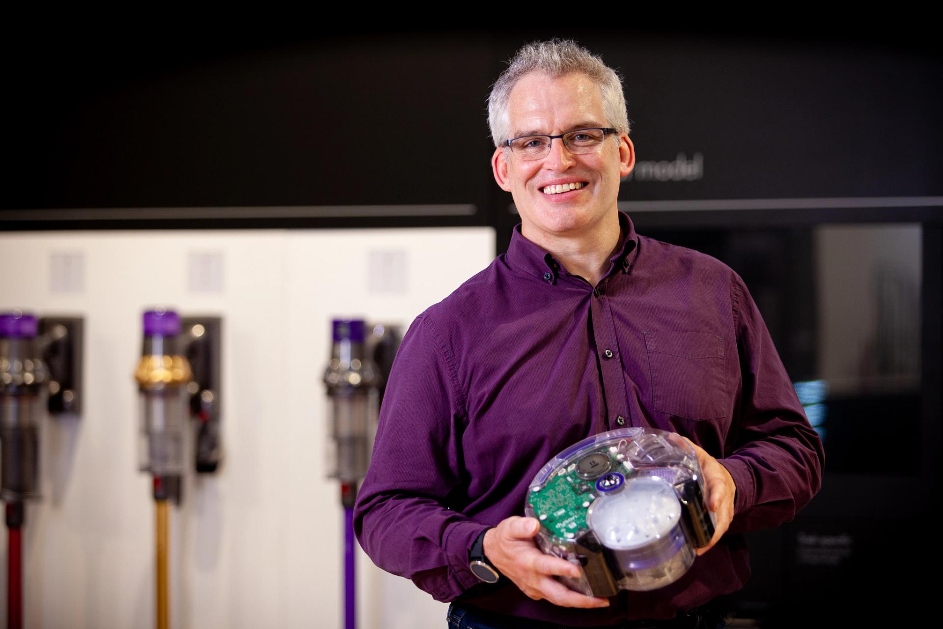 Mike Aldred with a robot vacuum prototype in front of Dyson vacuums.