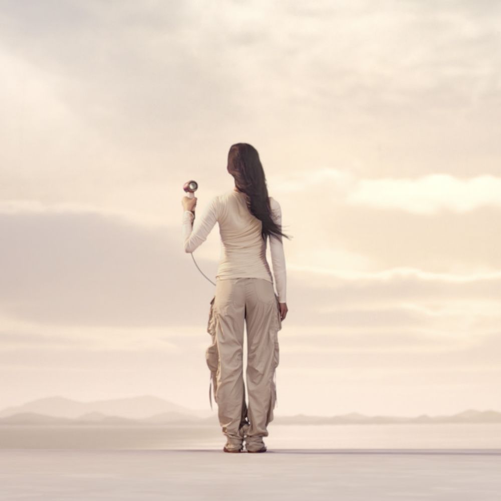 Model stands on a large flat landscape holding the Dyson Supersonic Travel hair dryer.