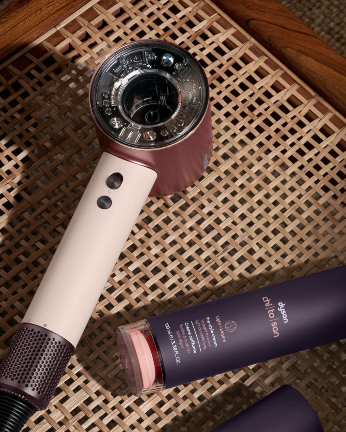 Secador de pelo Dyson Supersonic Nural sobre una silla de ratán con crema de prepeinado Dyson Chitosan. 