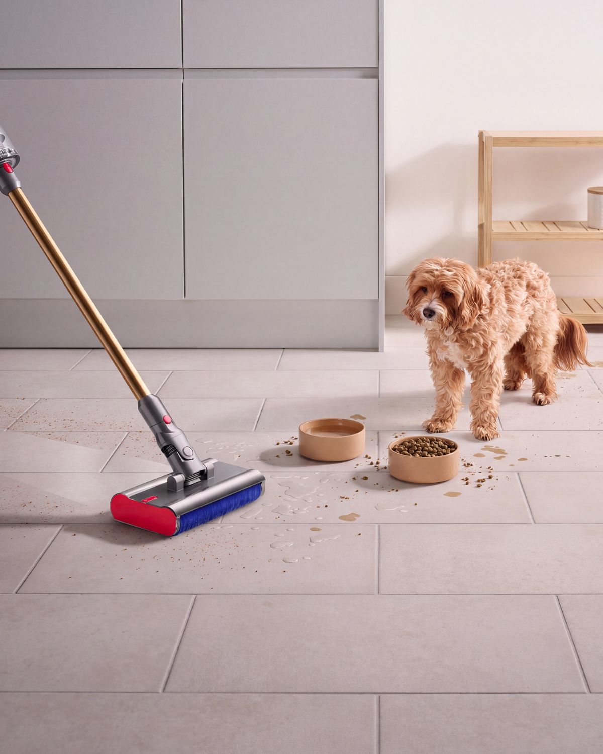 The Dyson V12s Detect Slim Submarine cleans the floor of a kitchen with a dog and dog food bowls in the background.