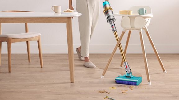 Un aspirateur Dyson ramasse les débris sur un sol dur. Un siège pour bébé se trouve à l'arrière-plan. 