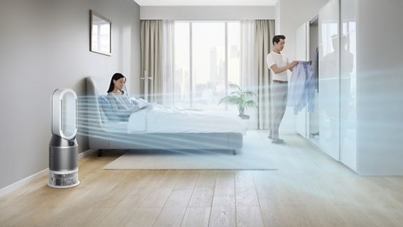 Femme étendue sur un lit pendant qu’un homme range une chemise dans la penderie.  Le Dyson Pure Humidify+Cool rafraîchit la pièce en arrière-plan.