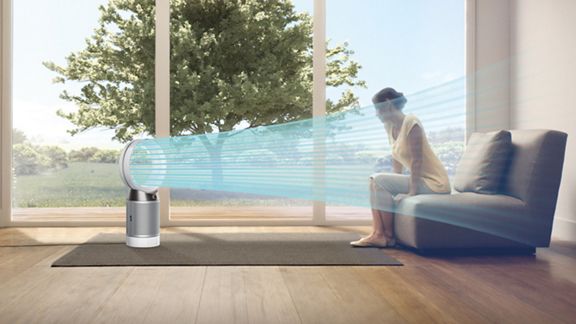 Une femme assise sur un canapé est entourée d’un jet d’air bleuté qui illustre l’air frais et purifié projeté par le ventilateur purificateur Dyson Cool. Dehors, il fait soleil. 