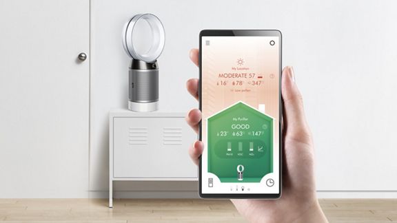 Un utilisateur tient un téléphone affichant l’application Dyson Link servant à contrôler le ventilateur purificateur Dyson Pure Cool positionné à l’arrière-plan. 