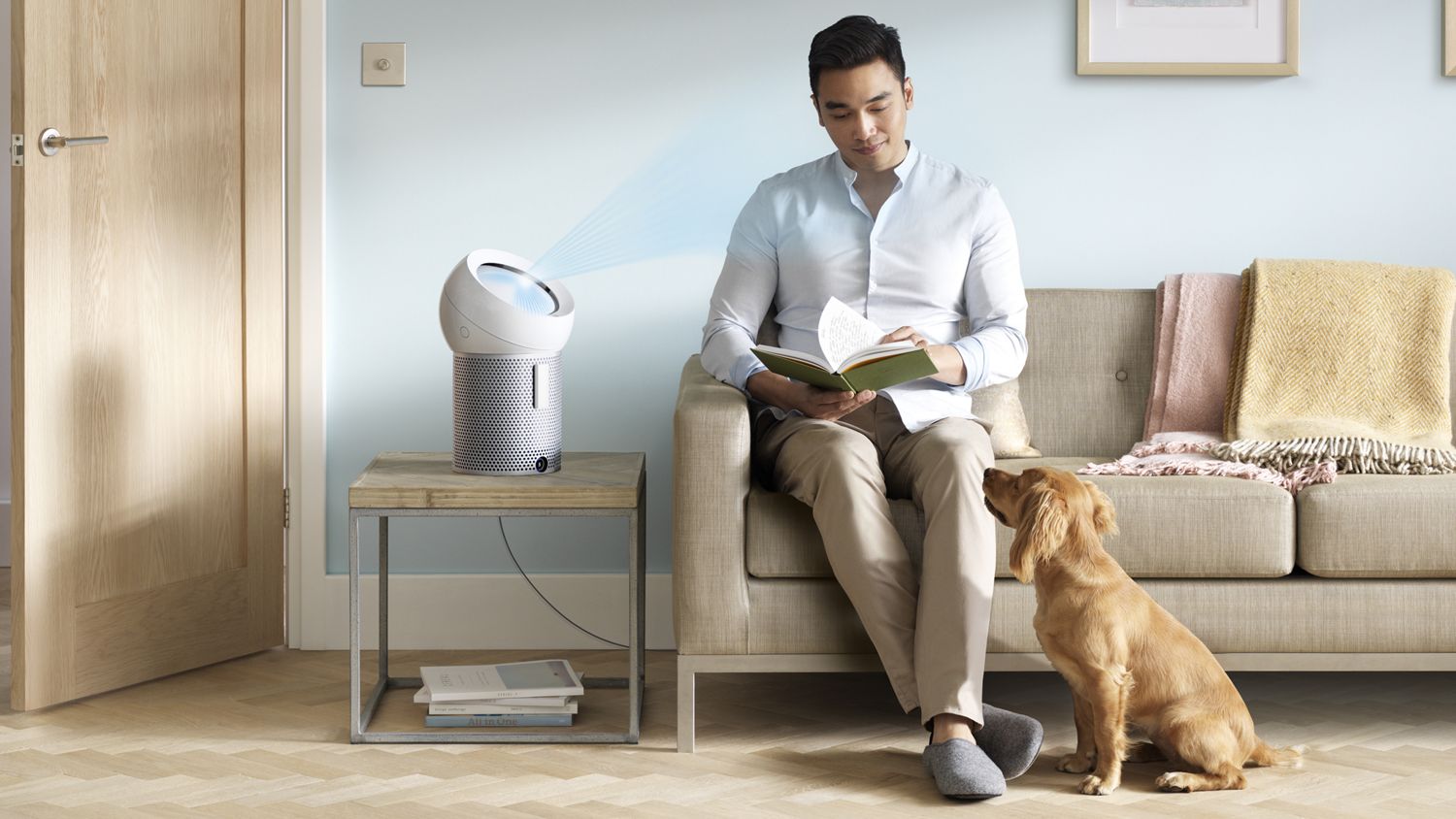 Un homme est assis sur un canapé avec un livre; un ventilateur purificateur personnel Dyson Pure Cool Me projette un flux d’air frais sur lui tandis qu’un chien s’assoit à ses pieds. 