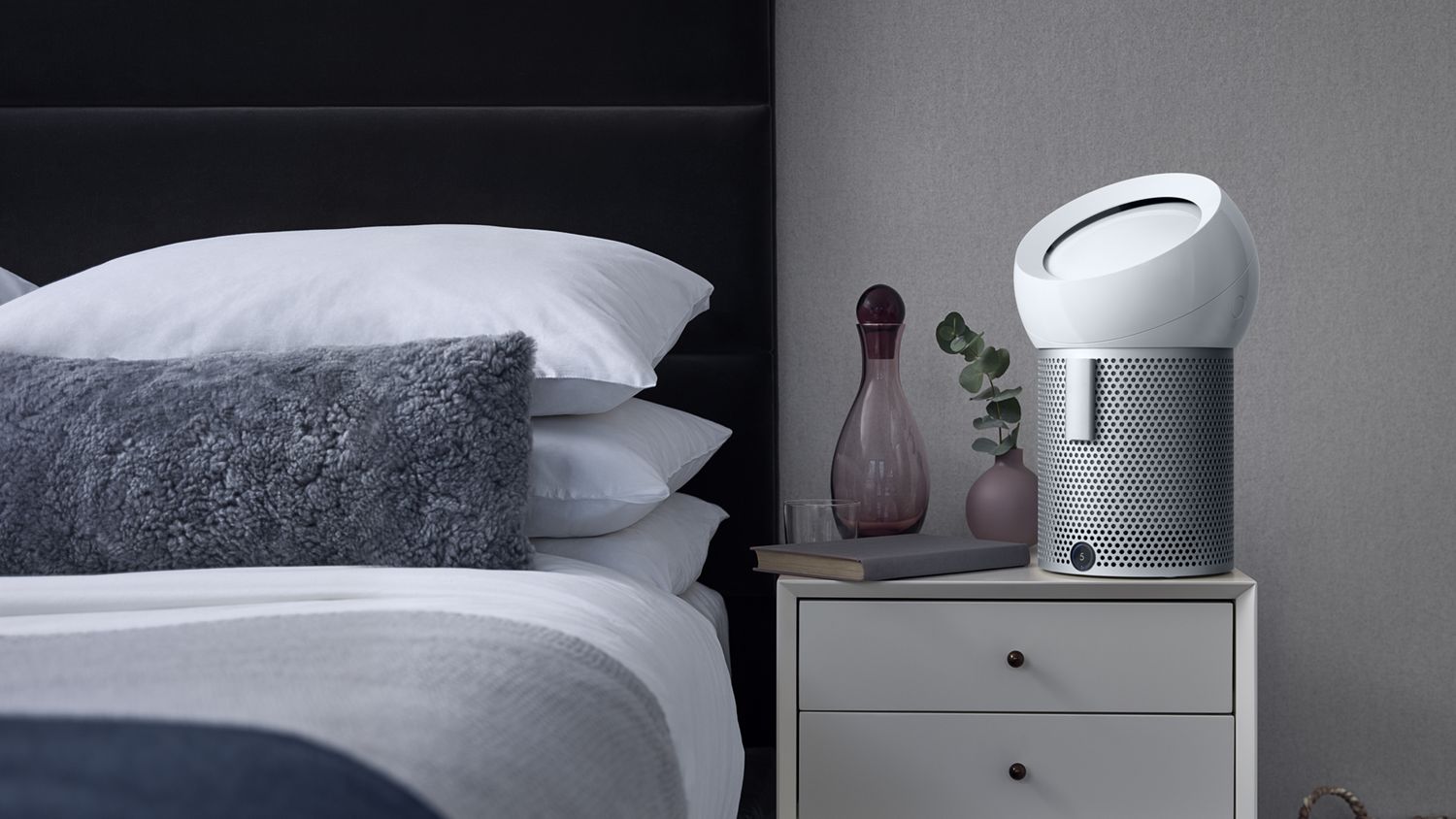 A Dyson Pure Cool Me personal purifier fan on a bedside table, angled towards the bed. 