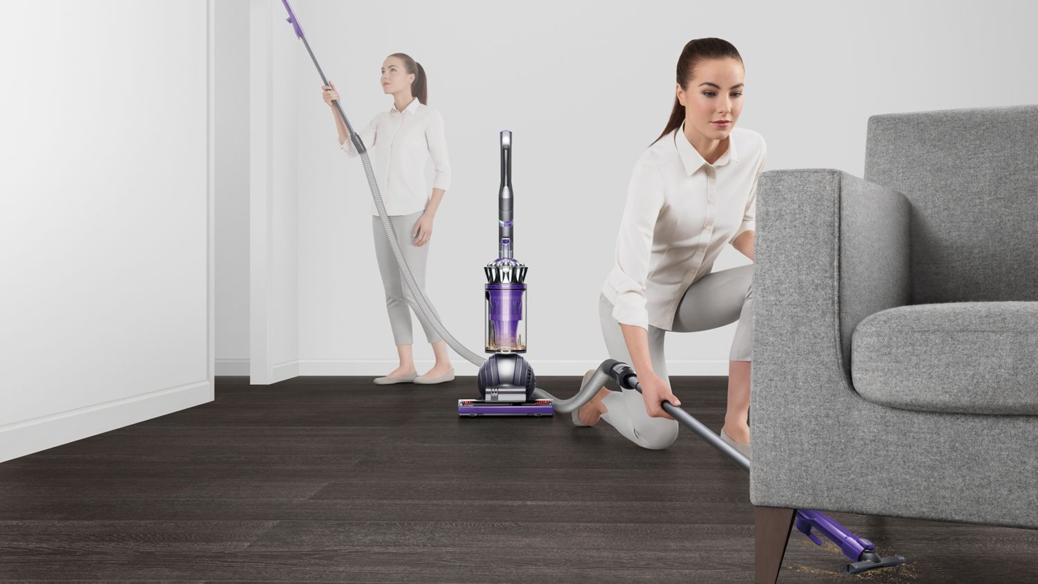 Dyson ball Animal 2 upright vacuum cleaner extendable hose being used to clean under a sofa. Woman in the background cleans a high area.
