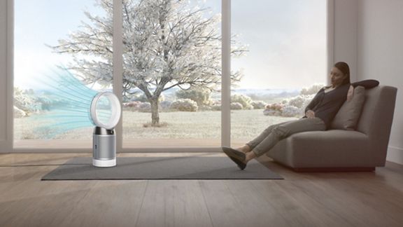 Eine Frau sitzt auf dem Sofa und ein blauer Luftstrom strömt durch die Hinterseite des Ventilators weg von der Frau. Es ist ein kalter Wintertag draußen. 