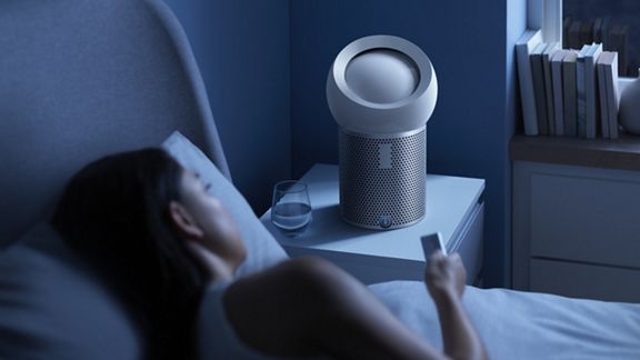 Eine Frau liegt nachts im Bett und hat eine Fernbedienung in der Hand, um ihren Dyson Pure Cool Me™ Luftreiniger für den persönlichen Gebrauch , der auf dem Nachttisch neben ihr steht, zu steuern. 
