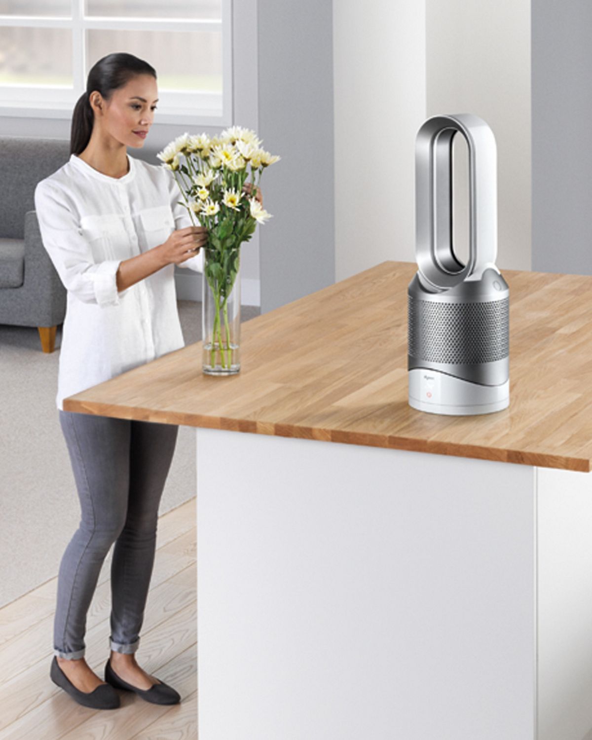 Um purificador termoventilador Dyson Pure Hot + Cool no meio de uma cozinha e alguém a arranjar as flores e a limpar a superfície de trabalho.