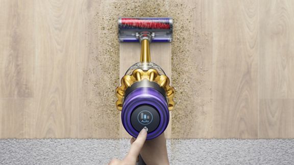Un utilisateur règle le mode de puissance d’un aspirateur sans fil Dyson V11 Absolute tandis que celui-ci ramasse les débris sur un sol dur et de la moquette.
