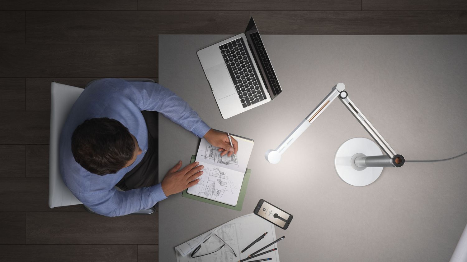 Eine Mann arbeitet an seinem Laptop, der auf einem Tisch steht. Die Dyson Solarcycle Arbeitsplatzleuchte beleuchtet seinen Arbeitsplatz. 