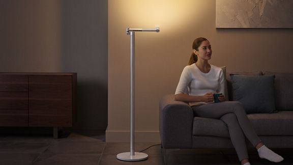 Eine Frau entspannt auf dem Sofa, während das Licht der Dyson Solarcycle Morph™ Leuchte an der Wand hinaufleuchtet und eine Umgebungsbeleuchtung erzeugt. 