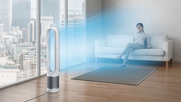 A woman sits on the sofa in a city apartment. A stream of blue, illustrating cool, purified air, projected from the Dyson Cool purifier fan onto her. 