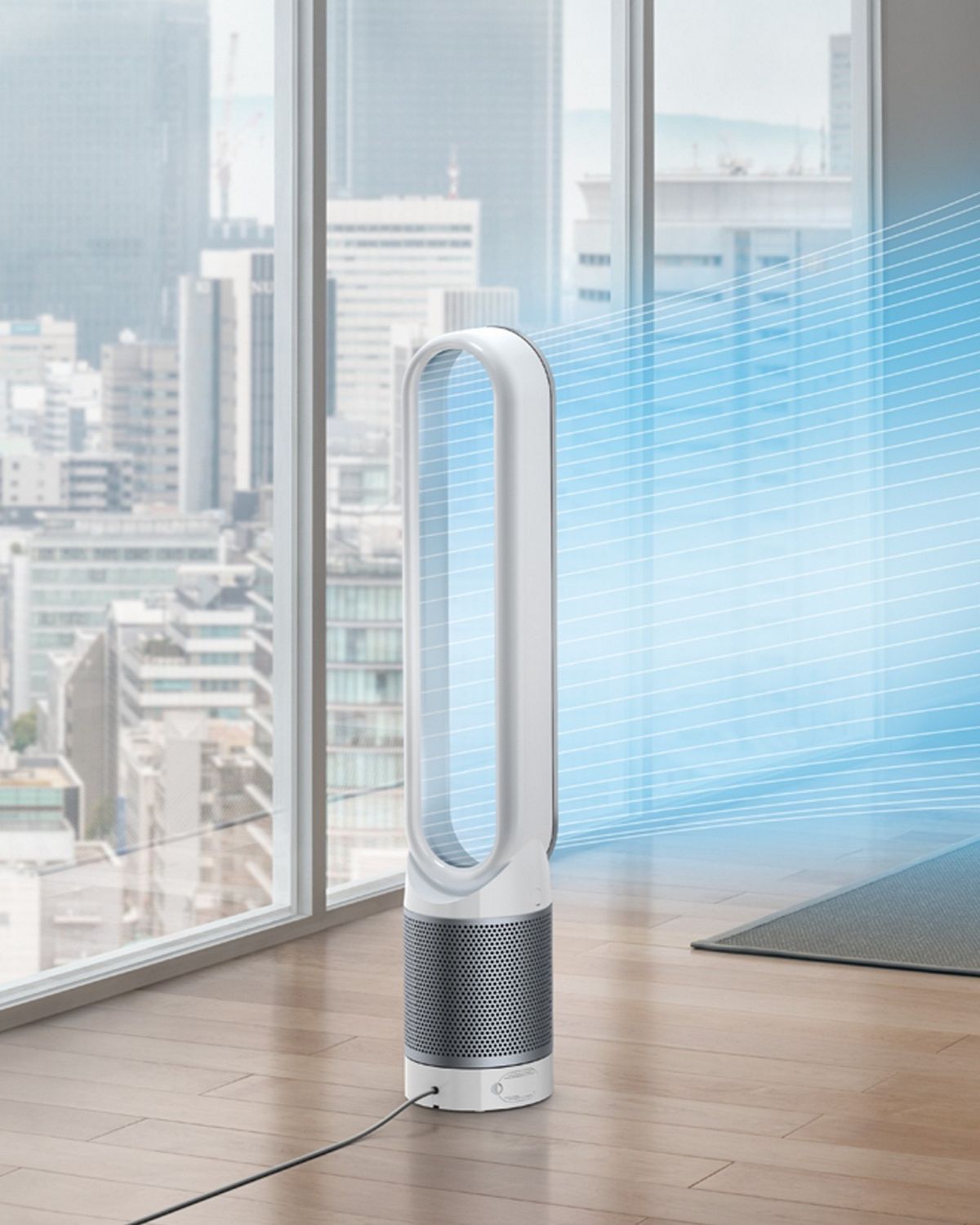 A woman sits on the sofa in a city apartment. A stream of blue, illustrating cool, purified air, projected from the Dyson Cool purifier fan onto her. 
