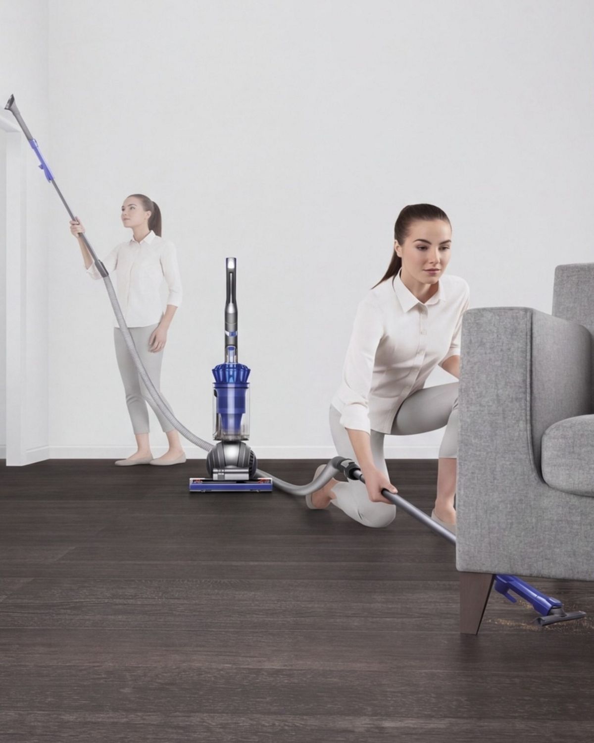 Dyson ball Total Clean upright vacuum cleaner extendable hose being used to clean under a sofa. Woman in the background cleans a high area.