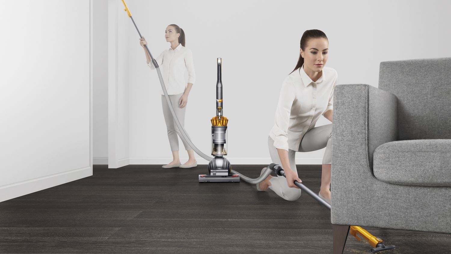 Dyson ball Multifloor 2 upright vacuum cleaner extendable hose being used to clean under a sofa. Woman in the background cleans a high area.
