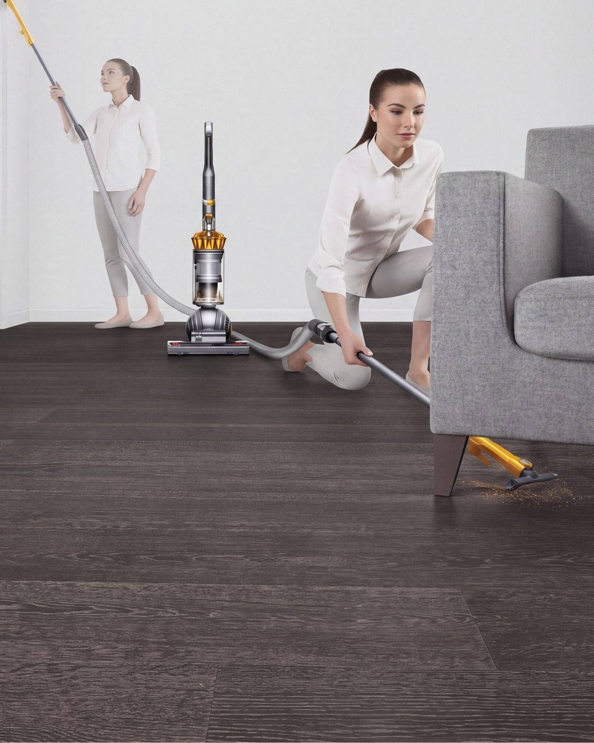 Dyson ball Multifloor 2 upright vacuum cleaner extendable hose being used to clean under a sofa. Woman in the background cleans a high area.