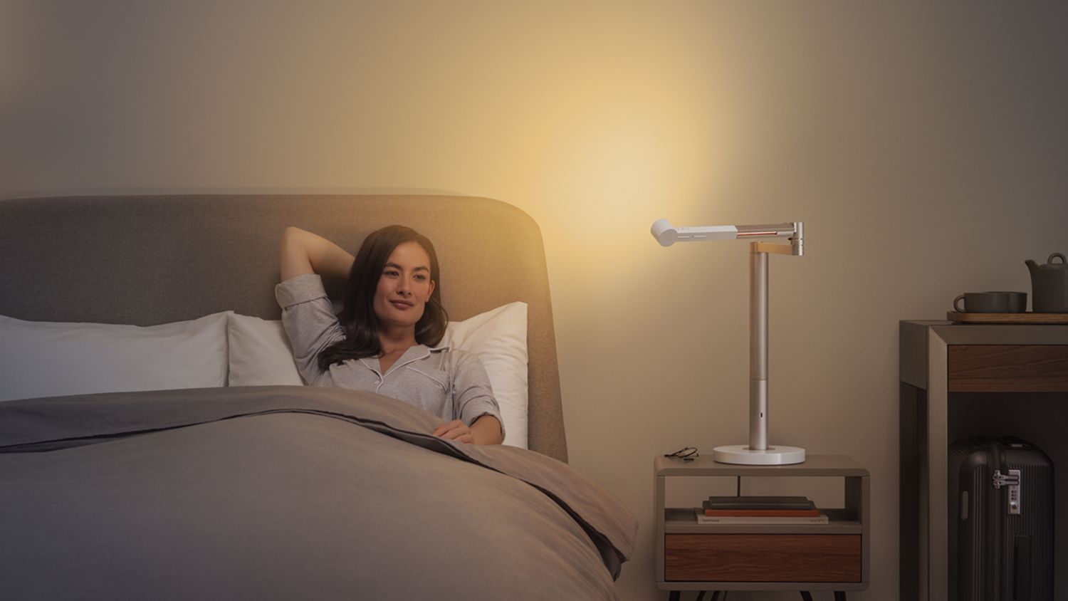 A woman sat in bed with the Dyson Lightcycle Morph light pointing at the wall beside her, creating indirect light in the room. 