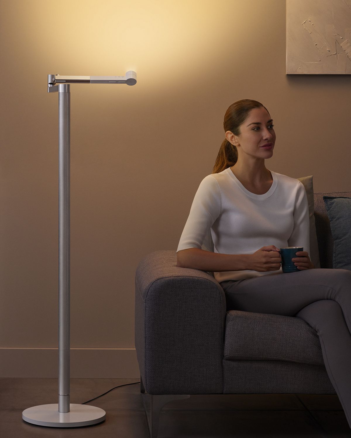 A woman relaxing on the sofa with the Dyson Lightcycle Morph light shining up at the wall above her, creating ambient light in the room. 
