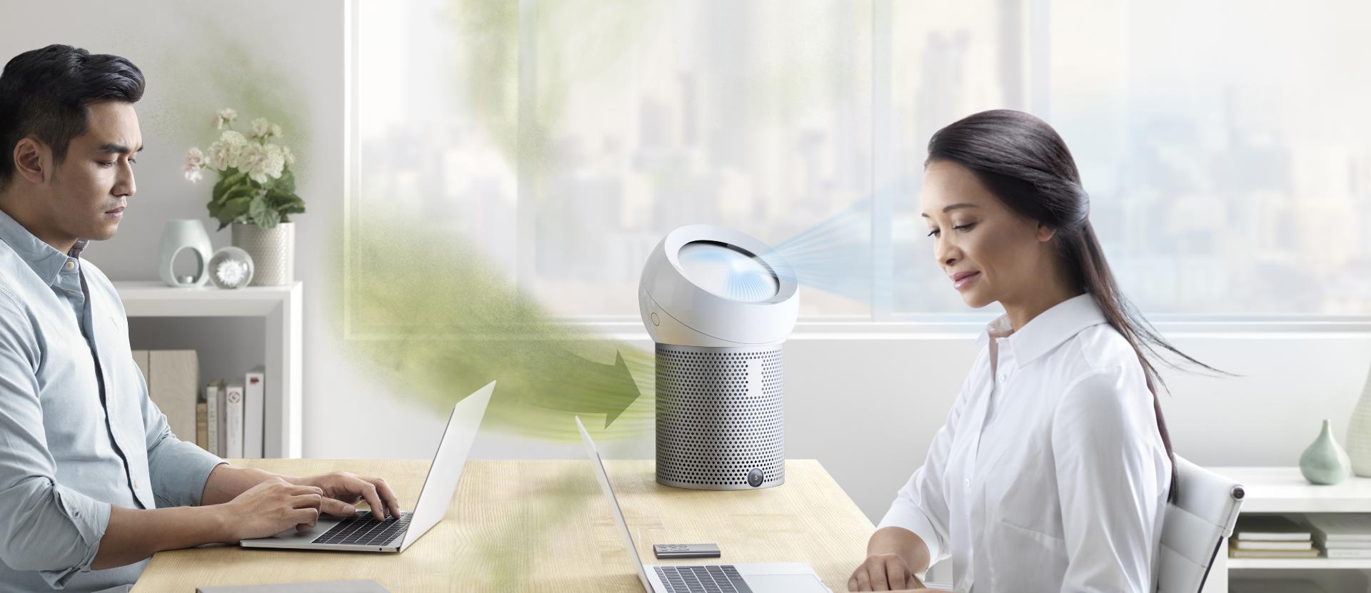 Mężczyzna i kobieta siedzą przy biurku, podczas gdy Dyson Pure Cool Me wchłania zanieczyszczone powietrze i rozprowadza oczyszczone powietrze