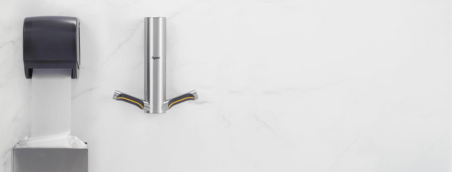 حائط مُثبَّت عليه مجفف الأيدي Dyson Airblade 9kJ بجوار جهاز توزيع مناشف ورقية كهربائي يَنسدل منه شريط طويل من المناشف الورقية متجه نحو سلة النفايات بالأسفل.