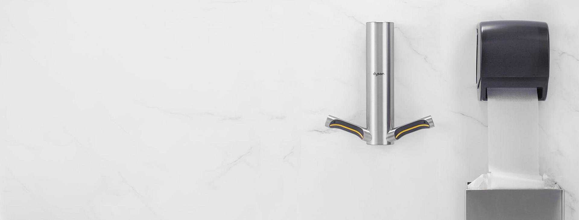 Dyson Airblade 9kJ hand dryer mounted beside a paper towel dispenser with sheet of paper towels going into trash can.