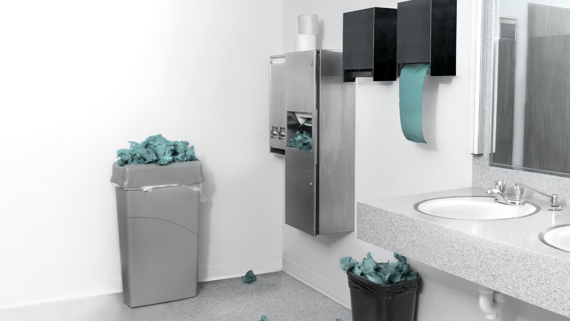 A public bathroom with paper towel dispensers and trash cans overflowing with paper towel waste. Some paper towels are balled up on floor.