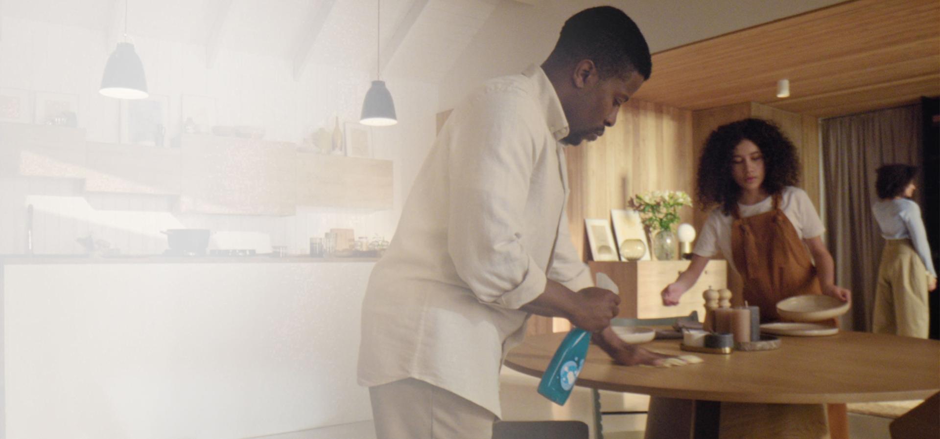 A man sprays cleaning product in a modern, open plan kitchen and living space just before meal time.