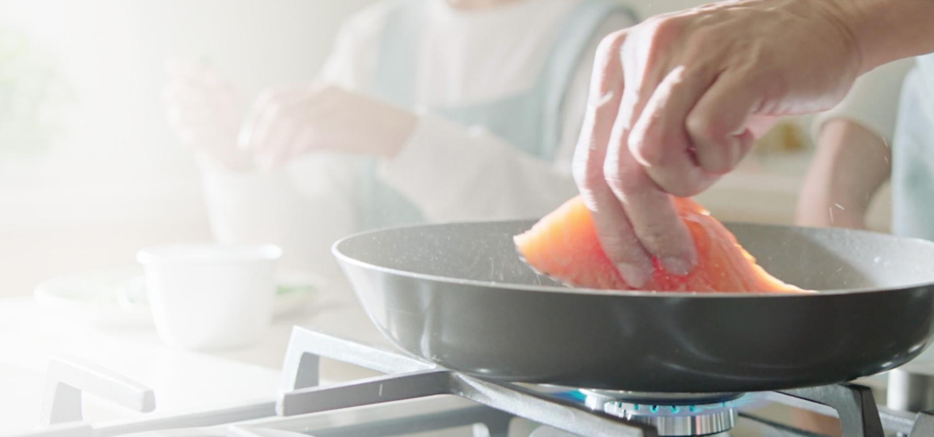 A close-up of a frying pan on a gas hob, cooking a piece of salmon.