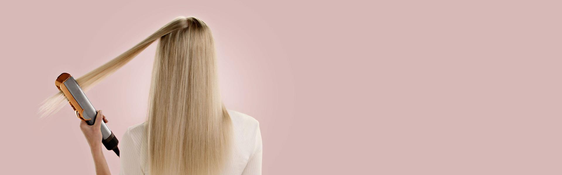 عارِضة شعرها أشقر طويل تُملِّس شعرها من الخلف باستخدام جهاز تمليس الشعر Dyson Airstrait.