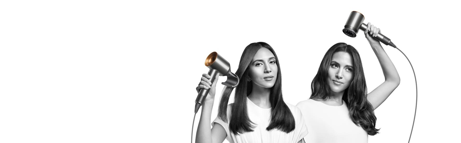 Dwie kobiety suszą włosy suszarką do włosów Dyson Supersonic. Kobieta po lewej używa końcówki do wygładzania puszących się włosów, aby wygładzić swoje proste, długie włosy, a kobieta po prawej używa końcówki suszącej delikatnym strumieniem.