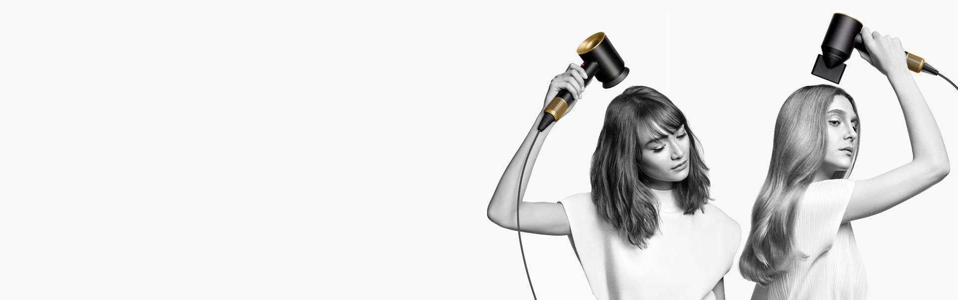 Two women dry their hair with the Dyson Supersonic hair dryer. The woman on the left uses the Flyaway attachment to smooth her straight long hair and the woman on the right uses the Diffuser attachment to shape her coily short hair.
