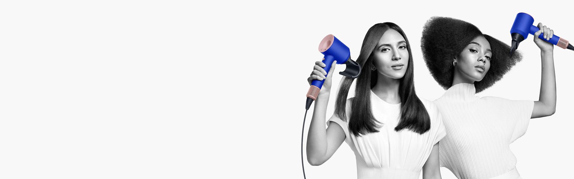 Two women dry their hair with the Dyson Supersonic hair dryer. The woman on the left uses the Flyaway attachment to smooth her straight long hair and the woman on the right uses the Diffuser attachment to shape her coily short hair.