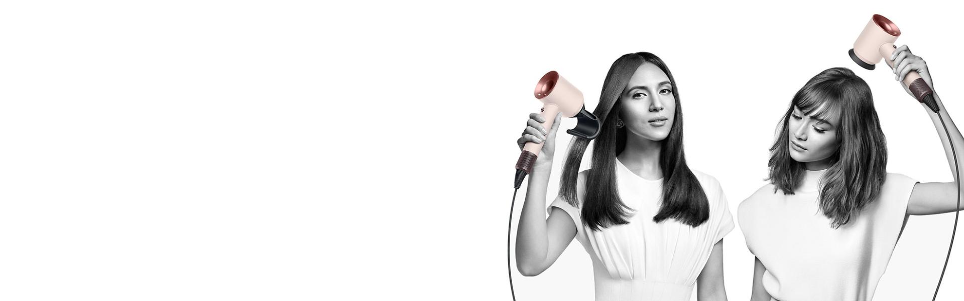 Two women dry their hair with the Dyson Supersonic hair dryer. The woman on the left uses the Flyaway attachment to smooth her straight long hair and the woman on the right uses the Diffuser attachment to shape her coily short hair.