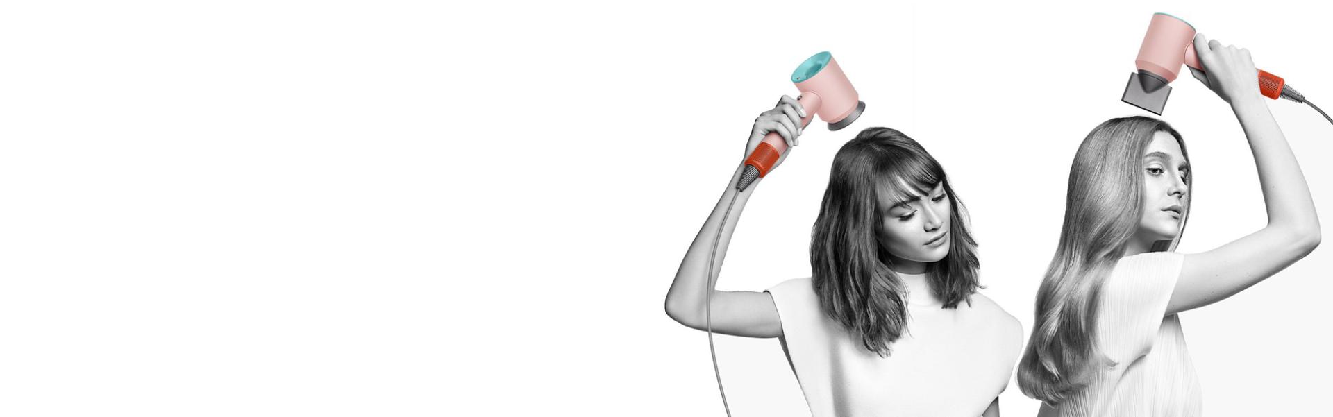 Two women dry their hair with the Dyson Supersonic hair dryer. The woman on the left uses the Flyaway attachment to smooth her straight long hair and the woman on the right uses the Diffuser attachment to shape her coily short hair.