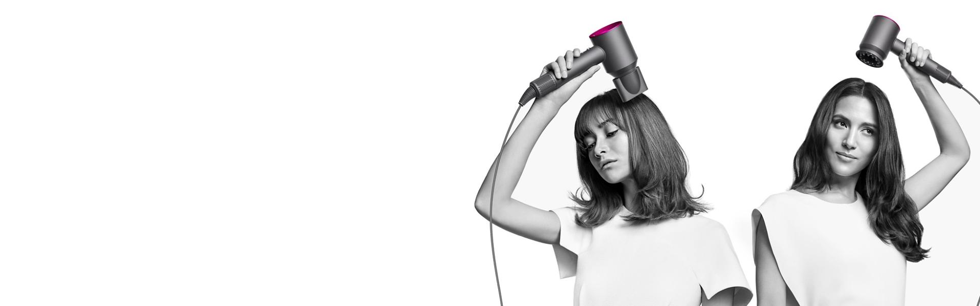Two women dry their hair with the Dyson Supersonic hair dryer. The woman on the left uses the Flyaway attachment to smooth her straight long hair and the woman on the right uses the Diffuser attachment to shape her coily short hair.