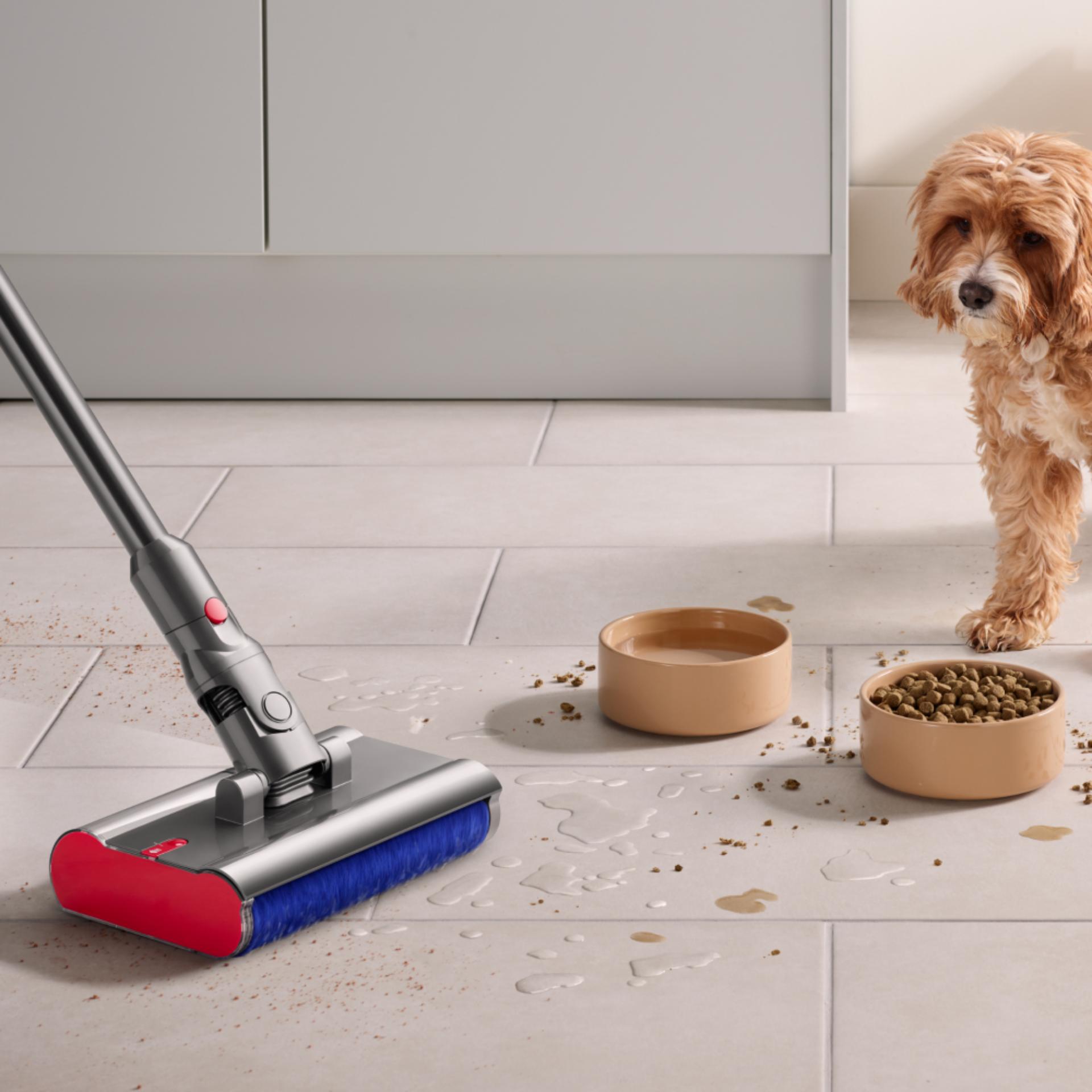 The Dyson V12s Detect Slim Submarine cleans the floor of a kitchen with a dog and dog food bowls in the background.