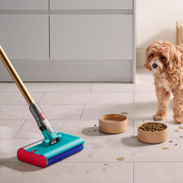 The Dyson V15s Detect Submarine cleans the floor of a kitchen with a dog and dog food bowls in the background.